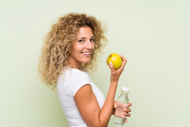 Junge blonde Frau mit dem lockigen Haar mit einem Apfel und mit einer Flasche Wasser