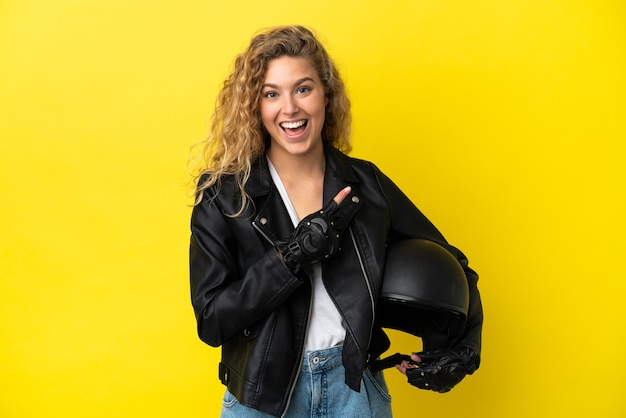 Junge blonde Frau mit einem Motorradhelm isoliert auf gelbem Hintergrund, der auf die Seite zeigt, um ein Produkt zu präsentieren