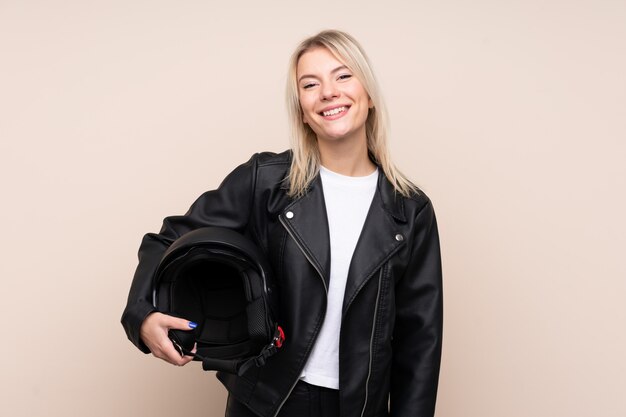 Junge blonde Frau mit einem Motorradhelm über isolierter Wand, die viel lächelt