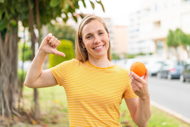 Junge blonde Frau mit einer Orange im Freien, stolz und selbstzufrieden