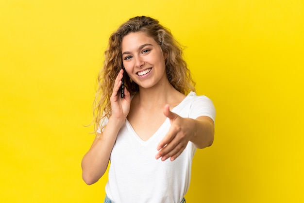 Junge blonde Frau mit Handy isoliert auf gelbem Hintergrund Händeschütteln für den Abschluss eines guten Deals