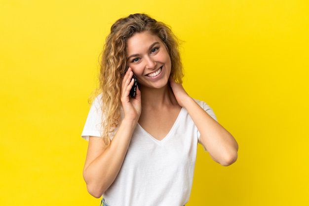 Junge blonde Frau mit Handy isoliert auf gelbem Hintergrund lachen