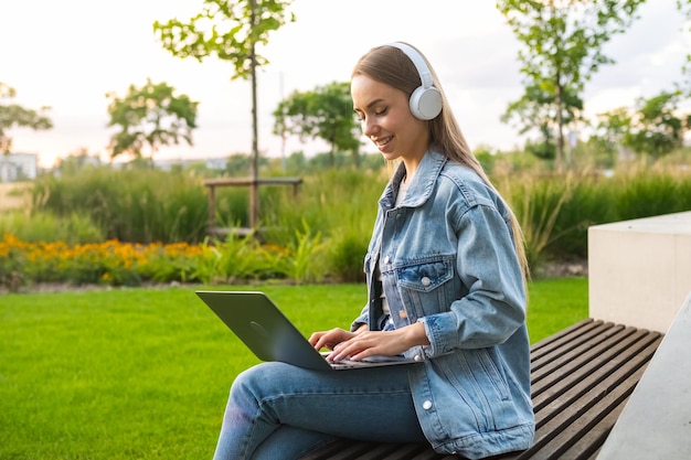 Junge blonde Frau mit Laptop auf dem Schoß schaut in die Kamera, sitzt am Abend auf einer Parkbank, Dame in Jeans
