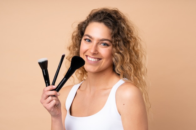 Junge blonde Frau mit lockigem Haar auf beige Wand, die Make-up-Pinsel hält