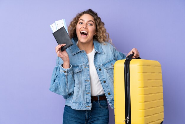 Junge blonde Frau mit lockigem Haar auf lila Wand im Urlaub mit Koffer und Pass und überrascht
