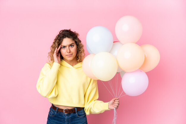 Junge blonde Frau mit lockigem Haar fängt viele Luftballons auf rosa Wand unglücklich und frustriert mit etwas.