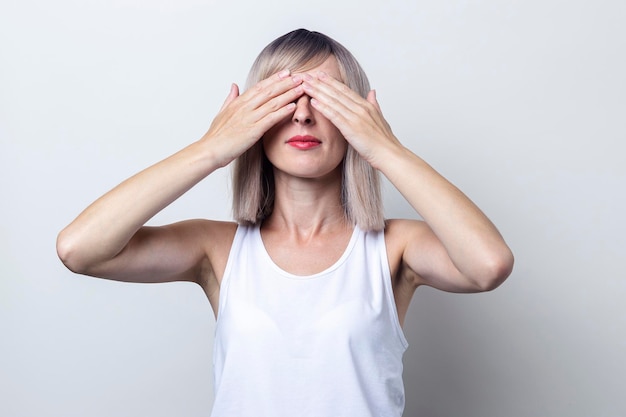 Junge blonde Frau mit seinen Händen, die ihre Augen auf einem hellen Hintergrund bedecken.