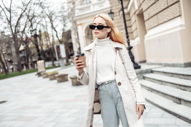 Junge blonde Frau mit Sonnenbrille und Trenchcoat hält unterwegs eine Tasse Kaffee in der Hand