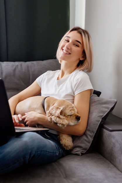 junge blonde Frau mit Spaniel sitzt an einem Laptop-Blogger und arbeitet im Internet-Home-Office