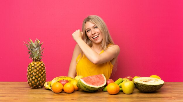 Junge blonde Frau mit vielen Früchten einen Sieg feiernd