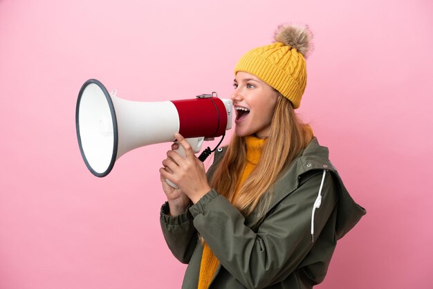 Junge blonde Frau mit Winterjacke isoliert auf rosa Hintergrund, die durch ein Megaphon schreit