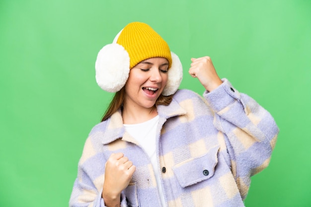 Junge blonde Frau mit Wintermuffs vor isoliertem Chroma-Key-Hintergrund feiert einen Sieg