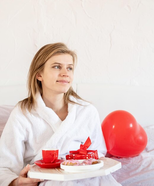 Junge blonde Frau sitzt im Bett mit Weihnachtsgeschenk und rotem Konfetti