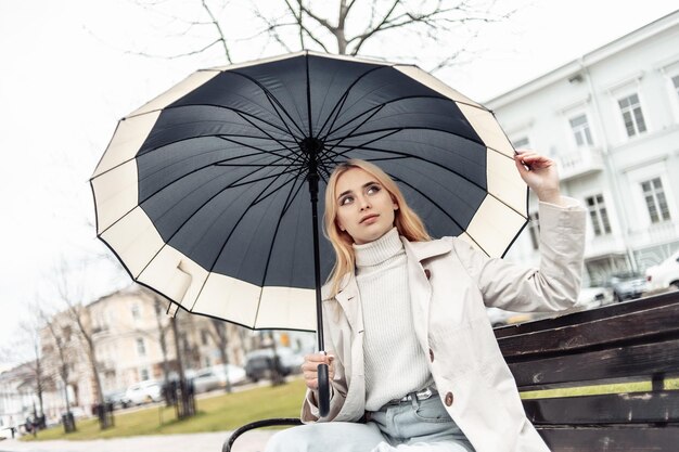 Junge blonde Frau sitzt mit Regenschirm auf einer Bank in der Stadt