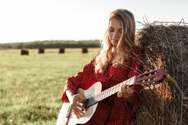 Junge blonde Frau spielt Gitarre in der Nähe eines Heuhaufens auf einem Feld im Sonnenuntergang