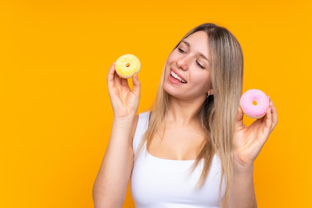 Junge blonde Frau über isolierte blaue Wand, die Donuts mit glücklichem Ausdruck hält