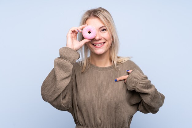 Junge blonde Frau über isolierte blaue Wand, die einen Donut und glücklich hält