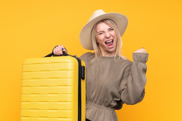 Junge blonde Frau über isolierte gelbe Wand im Urlaub mit Reisekoffer und Hut