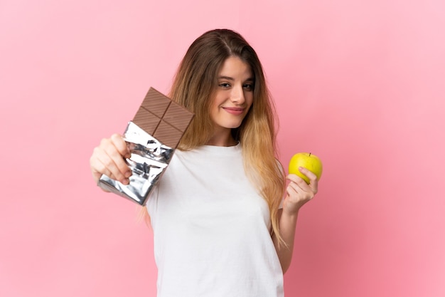 Junge blonde Frau über isoliertem Raum, der eine Schokoladentafel in einer Hand und einen Apfel in der anderen nimmt