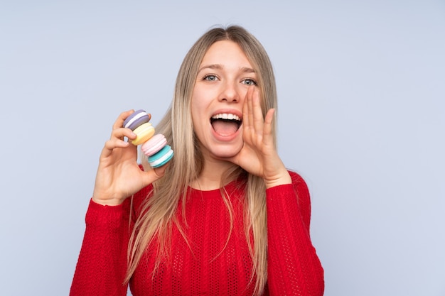 Junge blonde Frau über isolierter blauer Wand, die bunte französische Macarons hält und schreit