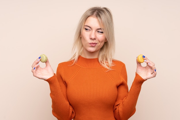 Junge blonde Frau über isolierter Wand, die bunte französische Macarons hält und denkt