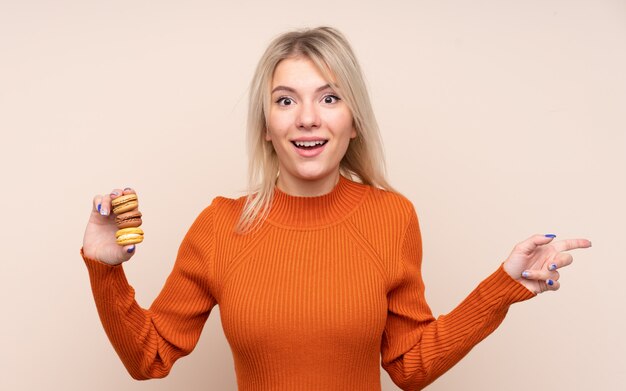 Junge blonde Frau über isolierter Wand, die bunte französische Macarons hält und Seite zeigt