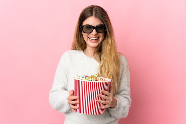 Junge blonde Frau über lokalisiertem Hintergrund mit 3D-Brille und hält einen großen Eimer Popcorn