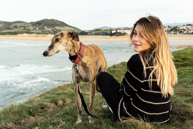 Junge blonde Frau und ihr Windhund lächelnd und genießen er Blick auf den Fluss zum Meer in Nordspanien