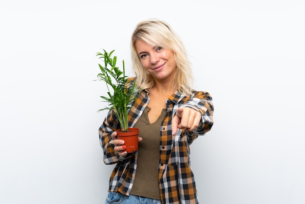 Junge blonde Gärtnerfrau, die eine Anlage über lokalisiertem Weiß hält, zeigt Finger auf Sie mit einem überzeugten Ausdruck