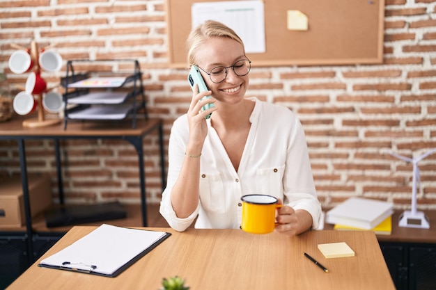 Junge blonde Geschäftsfrau spricht auf dem Smartphone und trinkt Kaffee im Büro