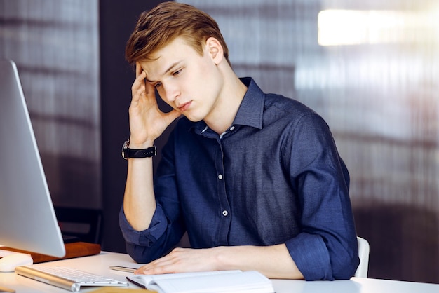 Junge blonde Geschäftsmann fühlt Stress am Arbeitsplatz in einem dunkeln Büro, Blendlicht im Hintergrund. Start-up-Geschäft bedeutet hartes Arbeiten und keine Zeit.