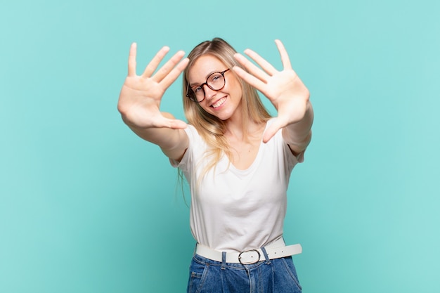 Junge blonde hübsche Frau, die lächelt und freundlich aussieht, die Nummer zehn oder zehn mit der Hand nach vorne zeigt und herunterzählt