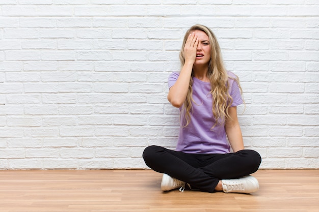 Junge blonde hübsche Frau, die schläfrig, gelangweilt und gähnend aussieht, mit Kopfschmerzen und einer Hand, die das halbe Gesicht bedeckt