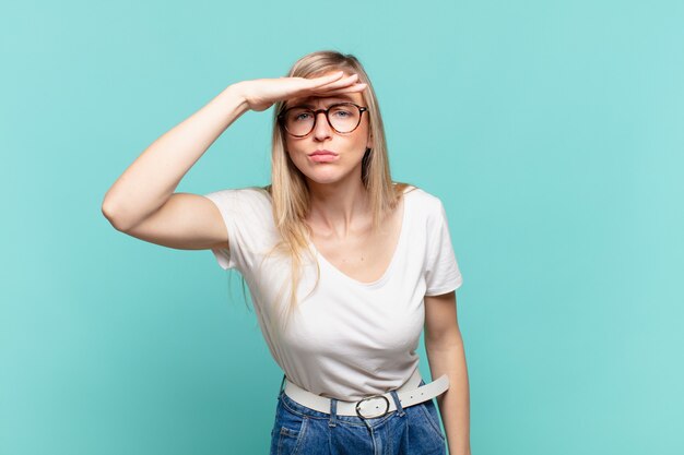 Junge blonde hübsche Frau, die verwirrt und erstaunt aussieht, mit der Hand über die Stirn, die weit weg schaut, zuschaut oder sucht