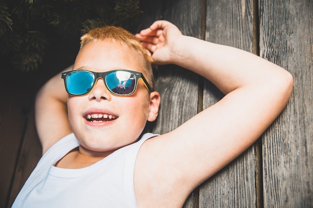 Junge blonde Junge mit Sonnenbrille liegt auf der Bank.