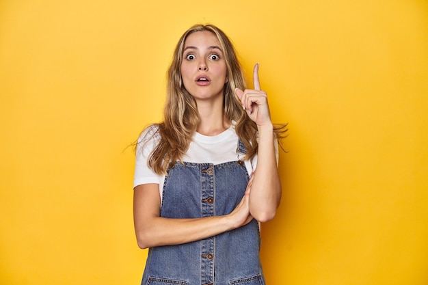 Junge blonde kaukasische Frau in Jeans-Overall, die auf einem gelben Hintergrund posiert und eine tolle Idee hat