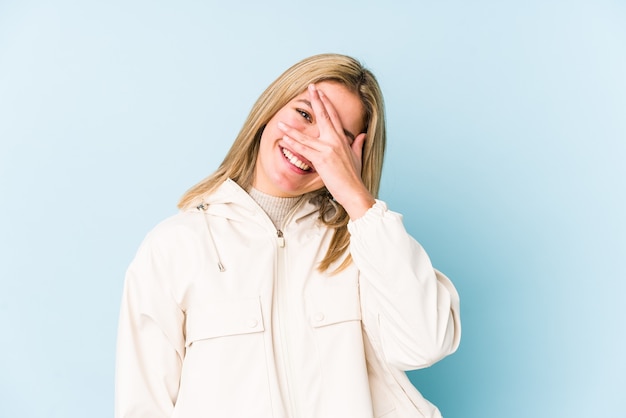 Junge blonde kaukasische Frau isolierte Blinzeln an der Kamera durch Finger, verlegenes bedeckendes Gesicht.