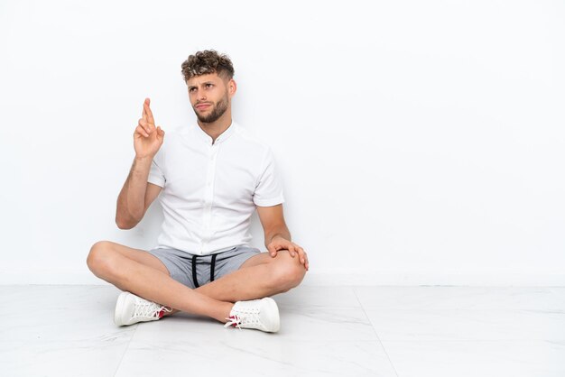Junge blonde Mann sitzt auf dem Boden isoliert auf weißem Hintergrund mit gekreuzten Fingern und wünscht das Beste