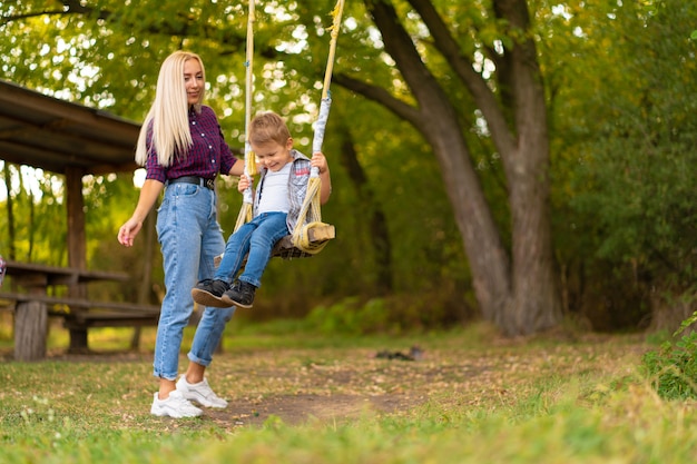 Junge blonde Mutter schüttelt ihren kleinen Sohn auf einer Schaukel in einem grünen Park. Glückliche Kindheit.