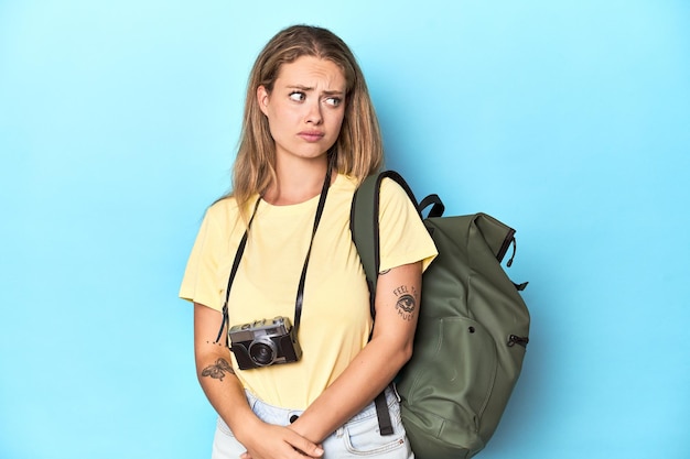 Junge blonde Reisende mit Kamera und Rucksack im Studio verwirrt fühlt sich zweifelhaft und unsicher