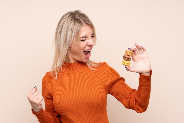 Junge blonde russische Frau über der lokalisierten Wand, die bunte französische macarons hält und einen Sieg feiert