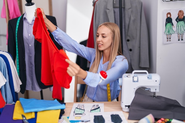 Foto junge blonde schneiderin lächelt selbstbewusst und hält t-shirt in der schneiderei