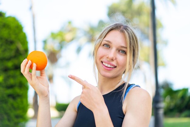 Junge blonde Sportlerin hält eine Orange im Freien und zeigt darauf