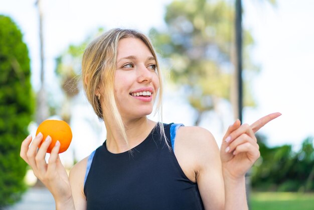 Foto junge blonde sportlerin hält eine orange im freien und zeigt zur seite, um ein produkt zu präsentieren