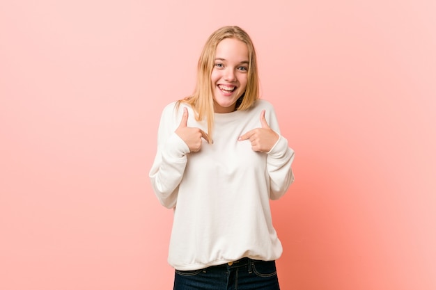 Junge blonde Teenagerfrau überrascht, mit dem Finger zeigend, breit lächelnd.