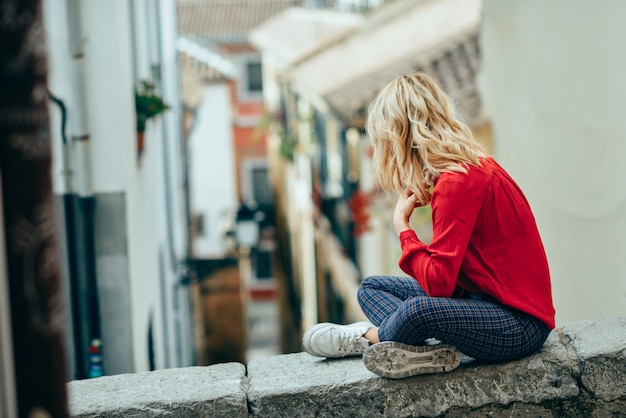 Junge blonde touristische Frau, die draußen sitzt, eine schöne schmale Straße schauend