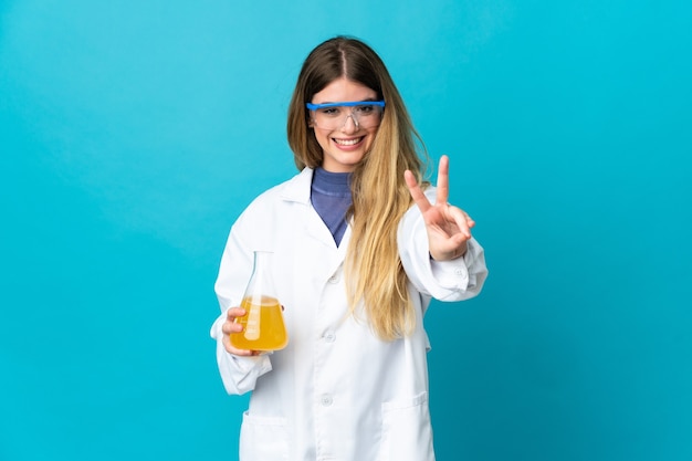Junge blonde wissenschaftliche Frau lokalisiert auf blauem Raum lächelnd und Siegeszeichen zeigend