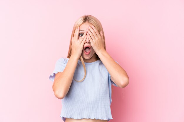 Junge Blondine auf rosa Wandblinzeln durch die Finger erschrocken und nervös.