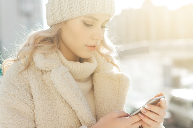 Junge blondine in der winterkleidung untersuchen den smartphone und das lächeln.