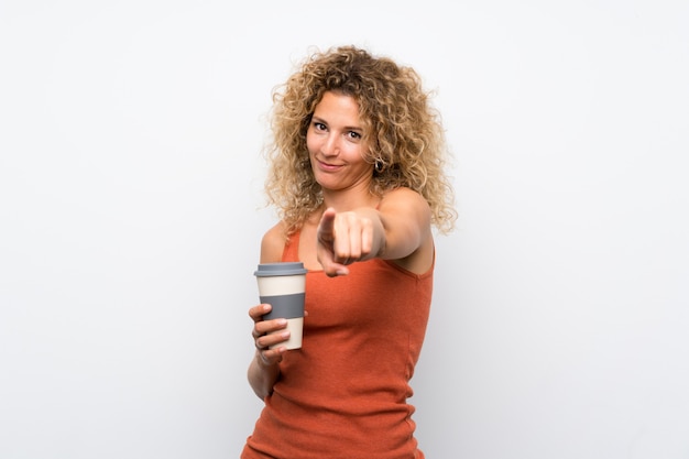 Junge Blondine mit dem gelockten Haar, das einen Mitnehmerkaffee hält, zeigen Finger auf Sie mit einem überzeugten Ausdruck